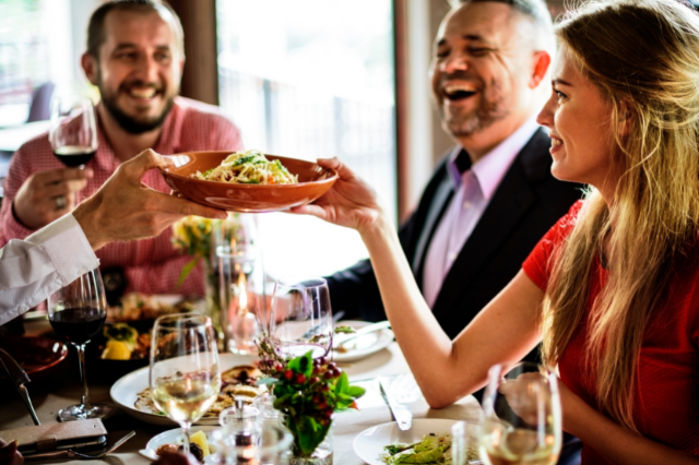 A woman passing a salad over to a man on the opposite side of a table in a restaurant. The table is full of food and glasses of wine. Two laughing men sit on the table next to the woman