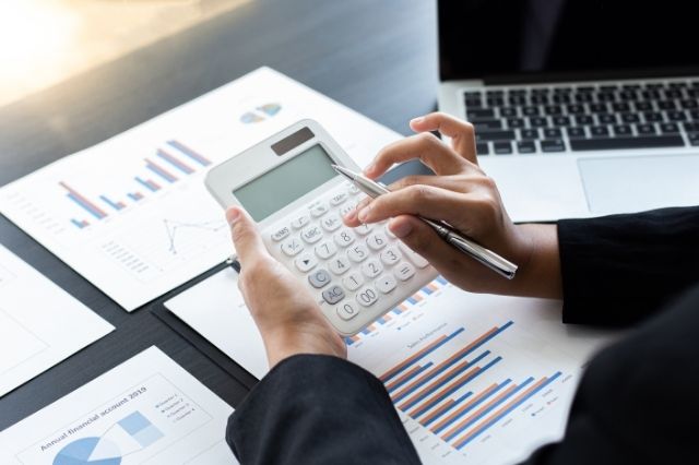 A person holding a small white calculator over a table with paper and a laptop. The paper has graphs and images on them. This person looks like they are an accountant