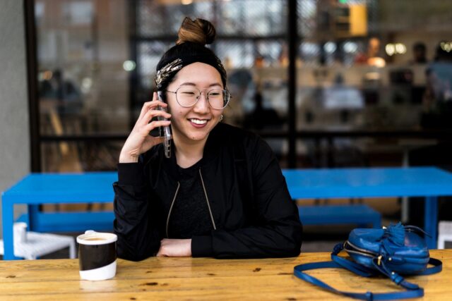 A woman with glasses and a bun is sitting at a table speaking on the phone and smiling. The table has a cup and her handbag on it.