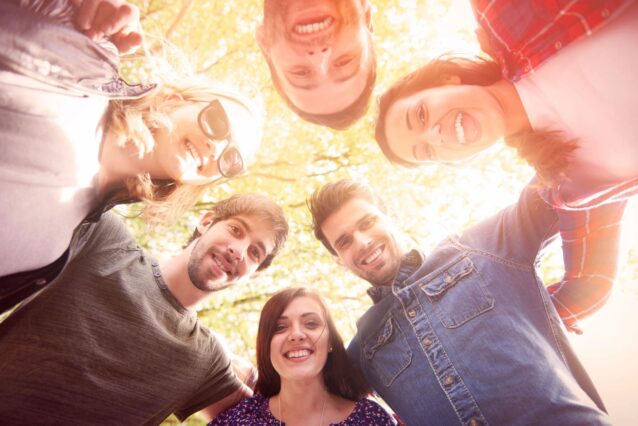 A group of men and women standing in a circle looking down at the camera looking as though they are working together. Camera is facing upwards as they all look down smiling below